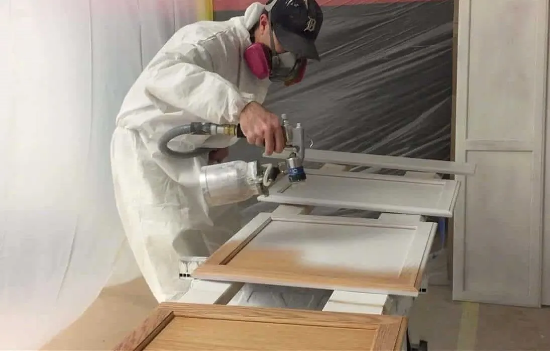 Painter spraying a cabinet door with protective clothing and masked surroundings